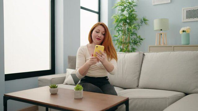 Young redhead woman make selfie by the smartphone sitting on sofa at home