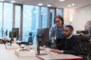 Young smiling woman explaining to serious African American coworker project strategy. Diverse startup coworkers students woman and man talking discussing working in modern office using computer.