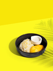 Thai Mango Sticky Rice, Southeast Asian dessert in black plate on yellow background with palm leaf shadow, clipping path.
