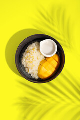 Thai Mango Sticky Rice, Southeast Asian dessert in black plate on yellow background with palm leaf shadow, clipping path.