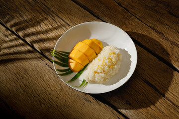 Thai Mango Sticky Rice, Southeast Asian dessert in white plate on wooden background with palm leaf shadow, clipping path.