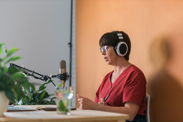 Portrait of mature woman wearing headphones and talking at online radio station - podcast and broadcast concept