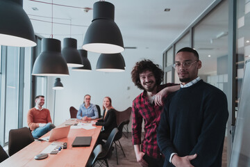 Portrait of two happy millennial male business owner in modern office. Two businessman smiling and looking at camera. Busy diverse team working in background. Leadership concept. Head shot.