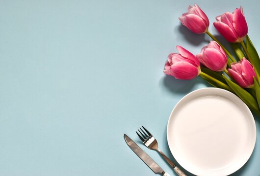 Festive creative table setting. Round plate and cutlery with tulip bouquet on blue background. Valentine's Day, Wedding Day, Birthday, Women's Day and Mother's Day. Flat lay. Copy space. Menu.