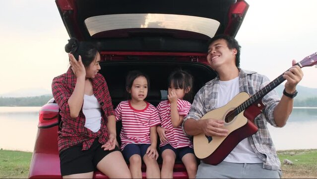 Happy Family Enjoying Road Trip On Summer Vacation. Mother And Child Sit In The Trunk Of The Car Singing Along With Dad Playing The Guitar. Holiday And Travel Family Concept.