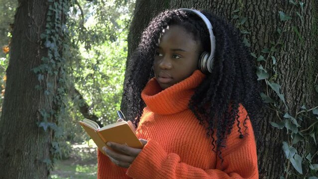 Black young woman using on white headphones while writing in her diary in a park