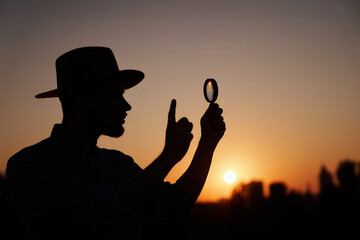 Silhouette of detective male in hat looking with magnifying glass raising index finger gesture. Man searching sales or discounts at sunset using loupe with sun on background and urban city view