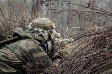 A man with a gun in camouflage and a panama hat is sitting in ambush in the grass, in the bushes. A soldier on a mission. The hunter takes aim.