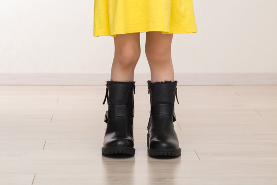 Little girl in yellow dress in mother big black leather boots on light floor at home room. Closeup. Front view.