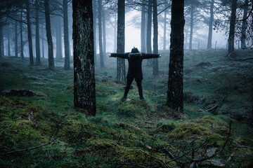 Man dressed in black with hood in mysterious cloudy forest. Person making cross shape with arms. Feeling of fear in the forest.