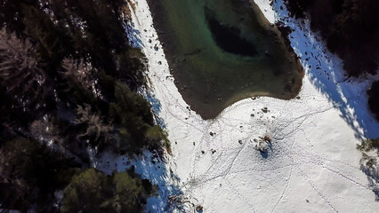 A drone shot of Green Lake in Austrian Alps. The lake shimmers with many shades of green and turquoise. Thick forest around it. Winter in the mountains. There is snow on the ground. Wandering