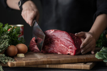 Dark and Moody. Chef in  holds a big knife and cuts into pieces raw meat on a brown wooden cutting...