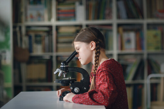 Cute caucasian girl schoolgirl with a pigtail with a microscope at home, natural science and research, children's cognitive interest