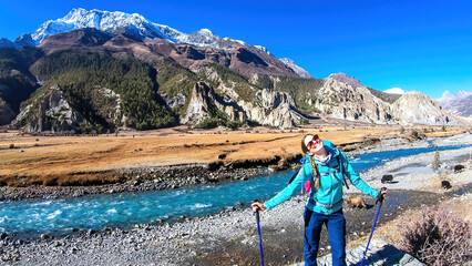 A woman with big hiking backpack admiring the view on the way to Manang, Annapurna Circuit Trek, Himalayas, Nepal. Some yaks are gazing on the meadow and crossing the river. Freedom and happiness