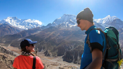 Fototapeta premium A couple trekking with panoramic view on Manang valley from Praken Gompa Nepal. High Himalayan ranges around. There is a small lake in the valley. Snow capped peaks of Annapurna Chain. Harsh landscape