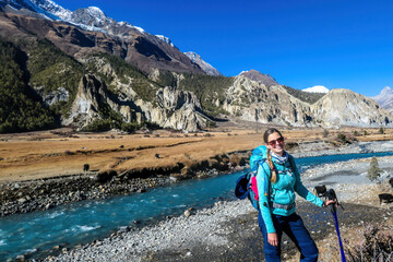 A woman with big hiking backpack admiring the view on the way to Manang, Annapurna Circuit Trek, Himalayas, Nepal. Some yaks are gazing on the meadow and crossing the river. Freedom and happiness
