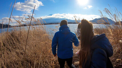 A caouple walking at the side of Kawaguchiko Lake, Japan with the view on Mt Fuji. They are enjoying the moment, having a lot of fun. Golden grass around them. The volcano surrounded by clouds.