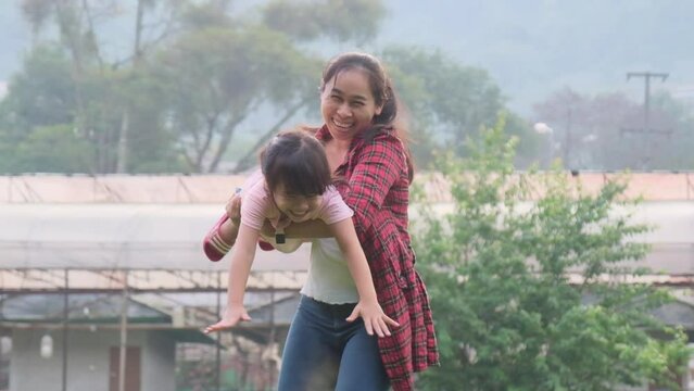 Happy Asian mother holds her daughter in her hands, little daughter raises her hand, imagining herself as a plane in the summer garden.