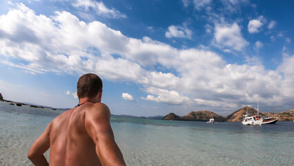 Fototapeta premium A man walking on a shallow sea water while taking selfies on Kelor Island in Komodo National Park, Indonesia. He is enjoying the moment, having fun while island hopping. There is a boat in the back
