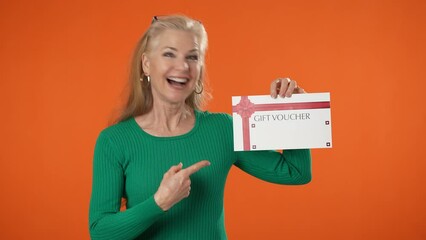 Happy mature blonde woman 40s 50s wearing green shirt hold gift certificate voucher winner gesture looking camera isolated on orange color background studio portrait.