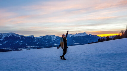 A girl walking on a snow covered lace, with high snow caped mountain range behind her in Schielfling am See, Austria. Sun is slowly setting behind the horizon. Cold winter afternoon.