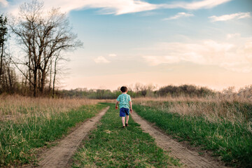 Naklejka na ściany i meble A young boy walks down a two track path between a grass field at sunset 