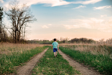 A young boy walks down a two track path between a grass field at sunset
