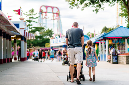 A father pushing a stroller and a daughter in a dress walk up the midway of an amusement park