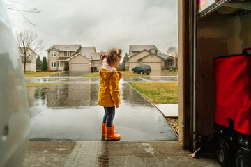 A young girl in a yellow rain jacket and orange rainboots stands in front of a wet driveway 