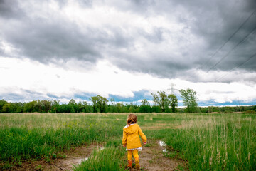 A young girl in a yellow raincoat and orange rainboots stands on a wet dirt path surrounded by wild grasses