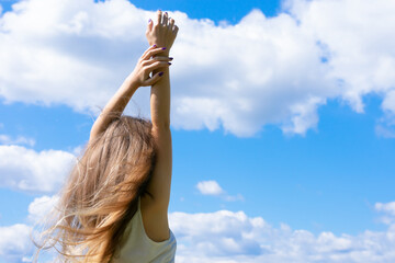 Blond haired young woman standing back on summer blue sky background with raised hands. Concept of...