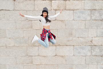 Full length portrait of a cheerful, casual Latina woman jumping isolated on grey stone wall.