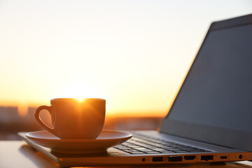 Coffee or tea cup on laptop against the window with sunshine. Cozy workplace in home office, inspiring view to morning city