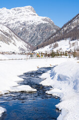 Fototapeta premium Scenic view of Livigno village in Sondrio province, Italy in winter. Popular skiing resort in European Alps. Snowcapped mountains, water stream and houses