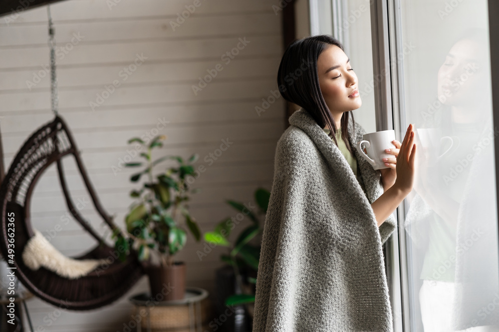 Wall mural young asian girl staying at home by the window, looking outside with nostalgic yearning face, holdin