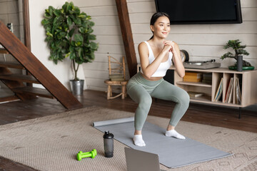 Young asian fitness girl doing squats workout at home, follow online sport gym instructor, standing on floor mat and smiling, exercising in living room