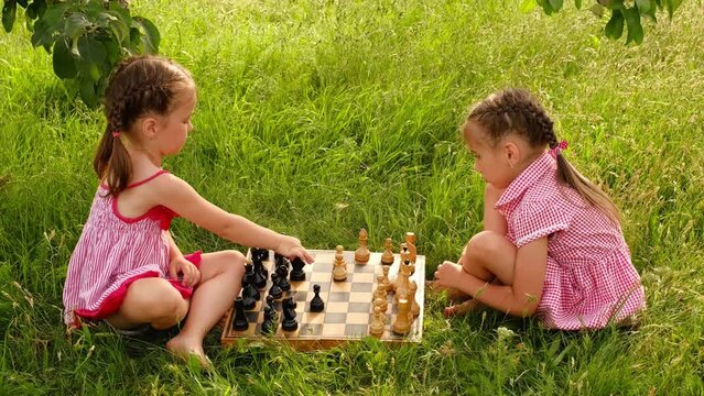 Two sisters are playing chess in the garden on the grass. An old wooden chessboard with pieces. Children's games on vacation. Rules of the game. Logical thinking. The girl makes a move with a knight