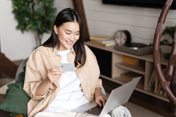 Smiling korean girl buying online from home, shopping on laptop and holding credit card
