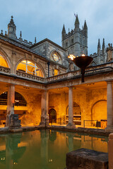 Obraz premium Bath town, UK. Evening sightseeing of restored in Victorian times ancient Roman Baths in the fire light of wall sconces. In the background visible tower of Abbey Church of Saint Peter and Saint Paul.