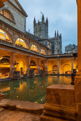 Bath town, UK. Evening sightseeing of restored in Victorian times ancient Roman Baths in the fire light of wall sconces. In the background visible tower of Abbey Church of Saint Peter and Saint Paul.