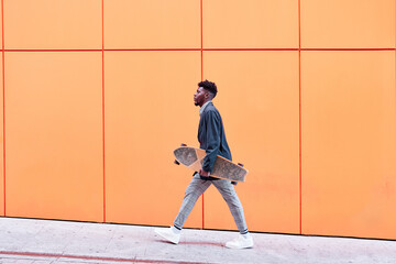 side view of a young man with blazer and skateboard walking down the street against an orange background. - Powered by Adobe
