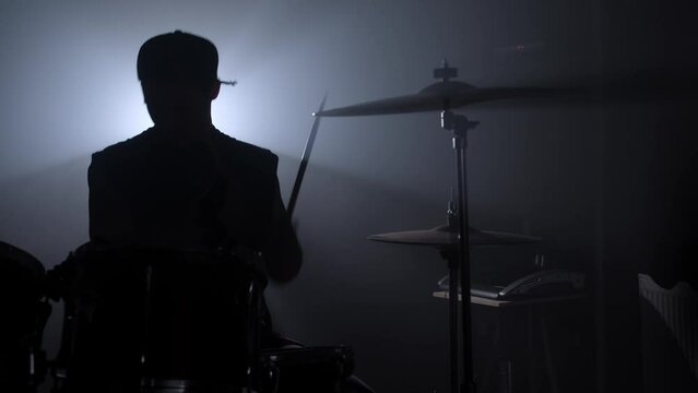 Silhouette drummer playing on drum kit on stage in a dark studio with smoke and neon lighting. Performance musical band. Slow motion.