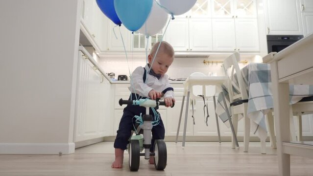 Kid on baby bicycle balance bike for toddlers with colorful balloons, 1 year old baby boy celebrating his first birthday at home. High quality 4k footage