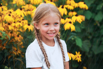 Close-up portrait of funny little girl with pigtails in yellow flowers in garden outdoor. Summertime