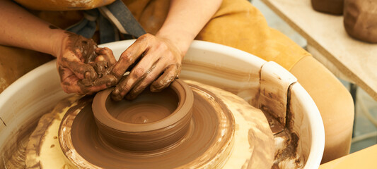 A Potter works with red clay on a Potter's wheel in the workshop..Women's hands create a pot. Girl sculpts in clay pot closeup. Modeling clay close-up. Warm photo atmosphere. 