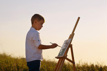 Little boy wearing white shirt standing in front of wooden easel and painting on canvas, rays of sunset on his face, sky on background. Side view artist working in open air. Concept of creativity