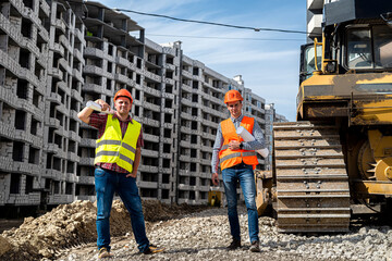 two young strong workers in uniform and helmets stand near a grader on a construction site