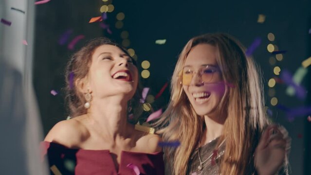 Close up couple of young multiracial girls wearing plush dresses enjoying glamorous party and having fun. Women dancing with falling confetti and sparkling or glowing sparklers or fireworks.