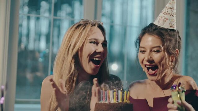 Close up portrait of young multiracial female friends blowing out candles on birthday cake and congratulating. Asian girl in party or birthday cap making wish at holiday party in apartment or house.