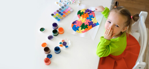 Girl drew a postcard for autism Day and sends an air kiss to all the children of the world. Gouache and watercolor paints in tubes and a palette on the table. A creative baby has drawn a puzzle-heart